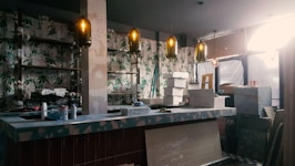 An indoor space under renovation, featuring a bar counter with various construction materials and tools scattered around. The walls are decorated with floral wallpaper. Multiple pendant lights hang from the ceiling, casting a warm glow. A ladder and several cardboard boxes are visible in the background.