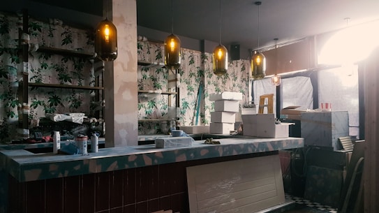 An indoor space under renovation, featuring a bar counter with various construction materials and tools scattered around. The walls are decorated with floral wallpaper. Multiple pendant lights hang from the ceiling, casting a warm glow. A ladder and several cardboard boxes are visible in the background.