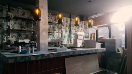 An indoor space under renovation, featuring a bar counter with various construction materials and tools scattered around. The walls are decorated with floral wallpaper. Multiple pendant lights hang from the ceiling, casting a warm glow. A ladder and several cardboard boxes are visible in the background.
