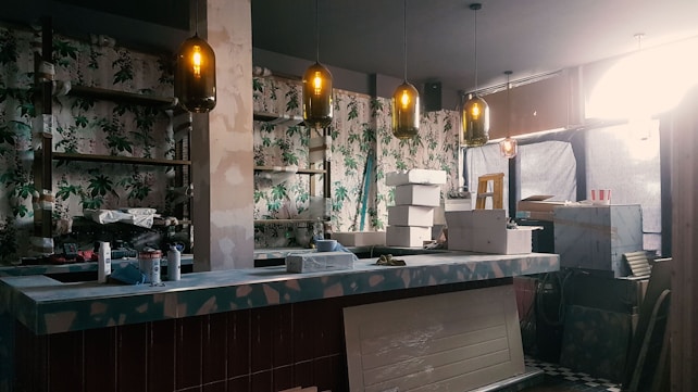 An indoor space under renovation, featuring a bar counter with various construction materials and tools scattered around. The walls are decorated with floral wallpaper. Multiple pendant lights hang from the ceiling, casting a warm glow. A ladder and several cardboard boxes are visible in the background.