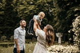 A family moment in a lush garden setting. A woman is joyfully lifting a young child into the air as a man smiles warmly beside them. The scene is surrounded by vibrant greenery and blossoming flowers.