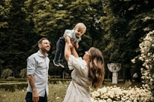 A family moment in a lush garden setting. A woman is joyfully lifting a young child into the air as a man smiles warmly beside them. The scene is surrounded by vibrant greenery and blossoming flowers.