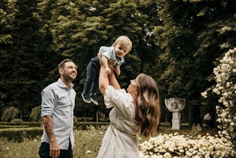 A family moment in a lush garden setting. A woman is joyfully lifting a young child into the air as a man smiles warmly beside them. The scene is surrounded by vibrant greenery and blossoming flowers.