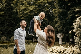 A family moment in a lush garden setting. A woman is joyfully lifting a young child into the air as a man smiles warmly beside them. The scene is surrounded by vibrant greenery and blossoming flowers.