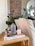 A cozy interior scene featuring a wooden table with stacked magazines and a sleek, modern diffuser. To the right, a light beige couch and to the left, green leafy plants add a touch of nature. In the background, an exposed brick wall provides texture, accompanied by a round mirror reflecting part of the room.