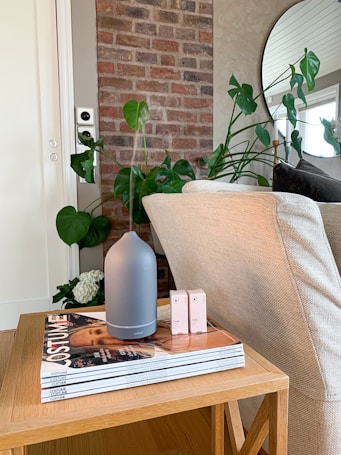 A cozy interior scene featuring a wooden table with stacked magazines and a sleek, modern diffuser. To the right, a light beige couch and to the left, green leafy plants add a touch of nature. In the background, an exposed brick wall provides texture, accompanied by a round mirror reflecting part of the room.