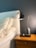 A bedside setup with a white upholstered headboard and cream pillows. A wooden nightstand is placed next to the bed, featuring a sleek black lamp and an essential oil diffuser emitting vapor. The background wall is painted in a dark color, complementing the overall decor.