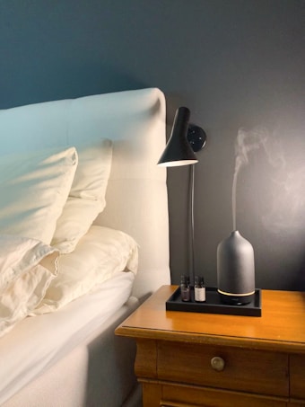 A bedside setup with a white upholstered headboard and cream pillows. A wooden nightstand is placed next to the bed, featuring a sleek black lamp and an essential oil diffuser emitting vapor. The background wall is painted in a dark color, complementing the overall decor.