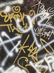 A wall covered in vibrant graffiti with swirling and angular designs in shades of yellow and white against a dark background. The graffiti includes various overlapping text and symbols, creating an intricate and urban texture.