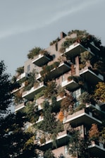 An urban residential building features numerous balconies filled with a lush array of greenery, including small trees and various plants. The architecture integrates nature into the living space, creating a vertical garden effect. The structure is bathed in natural light, which highlights the vibrant shades of green and orange foliage.