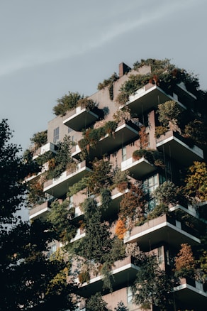 An urban residential building features numerous balconies filled with a lush array of greenery, including small trees and various plants. The architecture integrates nature into the living space, creating a vertical garden effect. The structure is bathed in natural light, which highlights the vibrant shades of green and orange foliage.