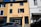 A two-story building with a yellow facade and blue shutters, featuring a real estate agency on the ground floor called Espaces Atypiques. A person is leaning out of an open window on the upper floor. There are parked cars in front of the building under a clear blue sky.