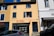 A two-story building with a yellow facade and blue shutters, featuring a real estate agency on the ground floor called Espaces Atypiques. A person is leaning out of an open window on the upper floor. There are parked cars in front of the building under a clear blue sky.