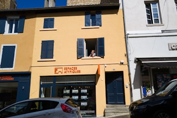 A two-story building with a yellow facade and blue shutters, featuring a real estate agency on the ground floor called Espaces Atypiques. A person is leaning out of an open window on the upper floor. There are parked cars in front of the building under a clear blue sky.