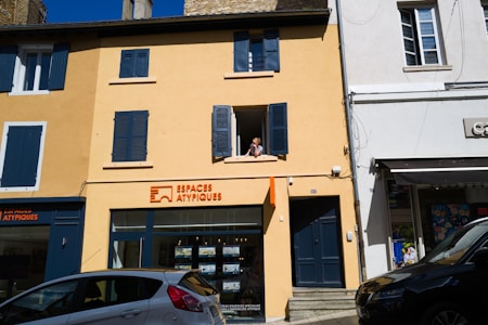 A two-story building with a yellow facade and blue shutters, featuring a real estate agency on the ground floor called Espaces Atypiques. A person is leaning out of an open window on the upper floor. There are parked cars in front of the building under a clear blue sky.