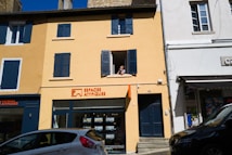 A two-story building with a yellow facade and blue shutters, featuring a real estate agency on the ground floor called Espaces Atypiques. A person is leaning out of an open window on the upper floor. There are parked cars in front of the building under a clear blue sky.