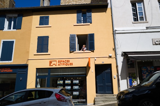 A two-story building with a yellow facade and blue shutters, featuring a real estate agency on the ground floor called Espaces Atypiques. A person is leaning out of an open window on the upper floor. There are parked cars in front of the building under a clear blue sky.