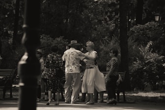 A group of elderly people is gathered in a park, engaging in social interaction. Some are dancing while others watch and converse. The setting is shaded by trees, creating a peaceful and intimate atmosphere.