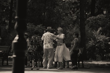 A group of elderly people is gathered in a park, engaging in social interaction. Some are dancing while others watch and converse. The setting is shaded by trees, creating a peaceful and intimate atmosphere.
