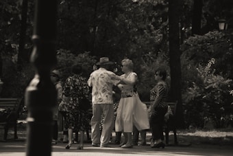 A group of elderly people is gathered in a park, engaging in social interaction. Some are dancing while others watch and converse. The setting is shaded by trees, creating a peaceful and intimate atmosphere.