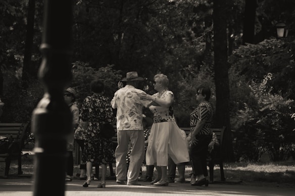 A group of elderly people is gathered in a park, engaging in social interaction. Some are dancing while others watch and converse. The setting is shaded by trees, creating a peaceful and intimate atmosphere.