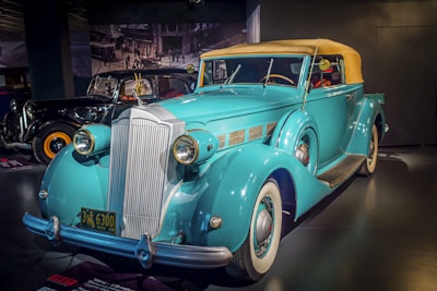 A classic blue convertible car with a beige soft top is prominently displayed in a showroom. The car features large round headlights, a prominent chrome grille, and white-walled tires. In the background, another vintage car is partially visible, suggesting a historic or museum setting.