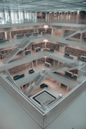 An interior of a spacious, multi-level modern library with bookshelves lining the walls. The library features clean lines and a minimalist design with white walls and staircases leading up multiple floors. The central atrium is open, with several seating areas scattered throughout.