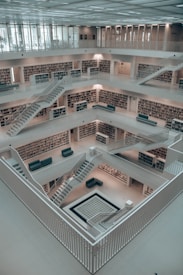 An interior of a spacious, multi-level modern library with bookshelves lining the walls. The library features clean lines and a minimalist design with white walls and staircases leading up multiple floors. The central atrium is open, with several seating areas scattered throughout.