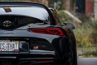 The image displays the rear view of a sleek black sports car, specifically showing its tail lights, rear bumper, and a Florida license plate. The car is parked near a blurred urban background.