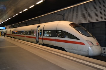 A modern high-speed train is stationed at an indoor platform, featuring a sleek design with a white and red color scheme. Several passengers can be seen standing near the train, and an electronic sign hangs above displaying information. The station has a contemporary architectural style with geometric patterns and strong lighting.