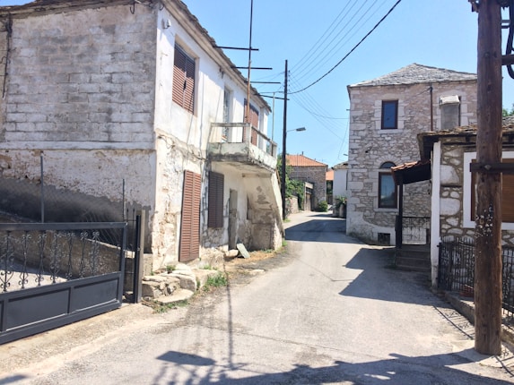 Narrow street in a rural village surrounded by old stone houses. The buildings display traditional architecture with wooden shutters and tiled roofs. Several electric wires crisscross the scene, and the road appears quiet and deserted.