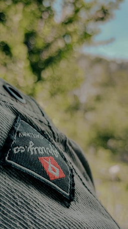A close-up view of a denim fabric featuring a woven patch with a logo. The patch shows a red diamond shape with a blue emblem inside, against a background of blurred greenery indicating an outdoor setting.