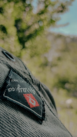 A close-up view of a denim fabric featuring a woven patch with a logo. The patch shows a red diamond shape with a blue emblem inside, against a background of blurred greenery indicating an outdoor setting.