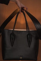 A black handbag with a sleek design is being held by a hand. The background is softly illuminated, showcasing the bag's polished texture and sturdy handles. The lighting creates a warm ambiance highlighting the bag.