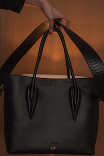 A black handbag with a sleek design is being held by a hand. The background is softly illuminated, showcasing the bag's polished texture and sturdy handles. The lighting creates a warm ambiance highlighting the bag.