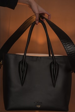 A black handbag with a sleek design is being held by a hand. The background is softly illuminated, showcasing the bag's polished texture and sturdy handles. The lighting creates a warm ambiance highlighting the bag.