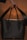 A black handbag with a sleek design is being held by a hand. The background is softly illuminated, showcasing the bag's polished texture and sturdy handles. The lighting creates a warm ambiance highlighting the bag.