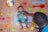 A baby in a striped green outfit is lying on a fish-themed inflatable ring surrounded by colorful plastic beach toys on a beige surface. An adult, partially visible, is gently holding the baby. There is a multicolored circular cake placed nearby.