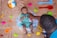 A baby in a striped green outfit is lying on a fish-themed inflatable ring surrounded by colorful plastic beach toys on a beige surface. An adult, partially visible, is gently holding the baby. There is a multicolored circular cake placed nearby.