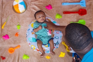 A baby in a striped green outfit is lying on a fish-themed inflatable ring surrounded by colorful plastic beach toys on a beige surface. An adult, partially visible, is gently holding the baby. There is a multicolored circular cake placed nearby.