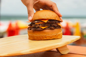 Hand holding a loaded burger stacked high with melted cheese and crispy bacon, with the beach skyline blurred in the background
