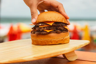 Hand holding a loaded burger stacked high with melted cheese and crispy bacon, with the beach skyline blurred in the background