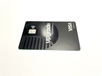 A credit card lies flat against a plain white background. The card features a sleek, minimalist design in dark and metallic shades. The word 'upgrade' is prominently featured along with text indicating Visa Signature.