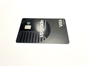 A credit card lies flat against a plain white background. The card features a sleek, minimalist design in dark and metallic shades. The word 'upgrade' is prominently featured along with text indicating Visa Signature.