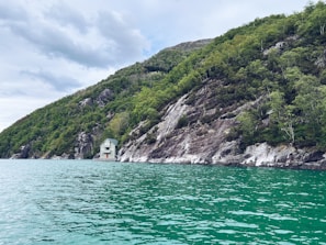 A small house is nestled at the base of a lush, green hillside with rocky outcrops. The house has a light exterior and contrasts with the natural surroundings. It is positioned right at the edge of a vibrant turquoise body of water, creating a peaceful and secluded ambiance. The sky is overcast with patches of cloud, suggesting a calm weather condition.