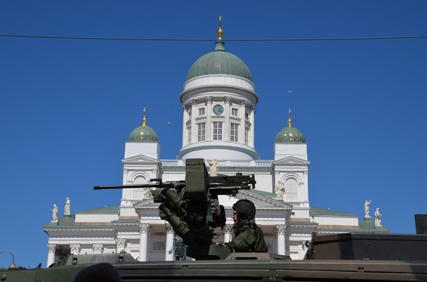 A large white cathedral with a green dome and gold accents is seen in the background, while in the foreground there is a military vehicle with a soldier operating a large mounted gun.