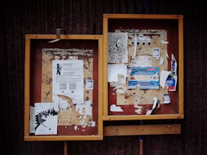 Two weathered bulletin boards with a variety of papers and flyers attached. Some flyers feature text with QR codes, while others contain artistic drawings and advertisements. The frames of the boards are wooden with a slightly worn appearance.