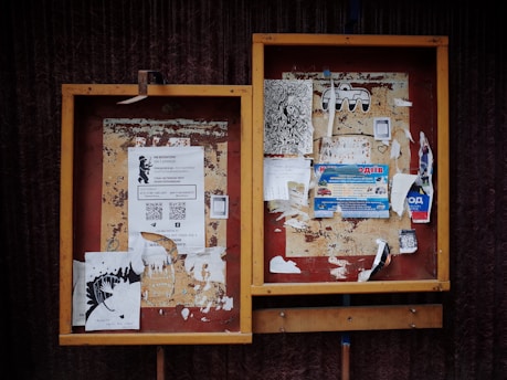 Two weathered bulletin boards with a variety of papers and flyers attached. Some flyers feature text with QR codes, while others contain artistic drawings and advertisements. The frames of the boards are wooden with a slightly worn appearance.