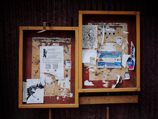 Two weathered bulletin boards with a variety of papers and flyers attached. Some flyers feature text with QR codes, while others contain artistic drawings and advertisements. The frames of the boards are wooden with a slightly worn appearance.