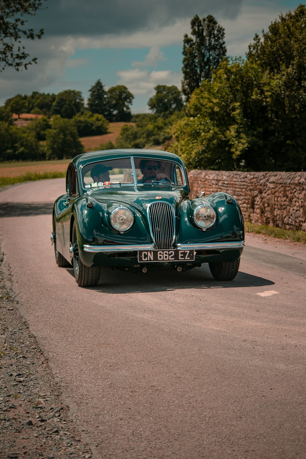 a green car driving down a road next to a stone wall